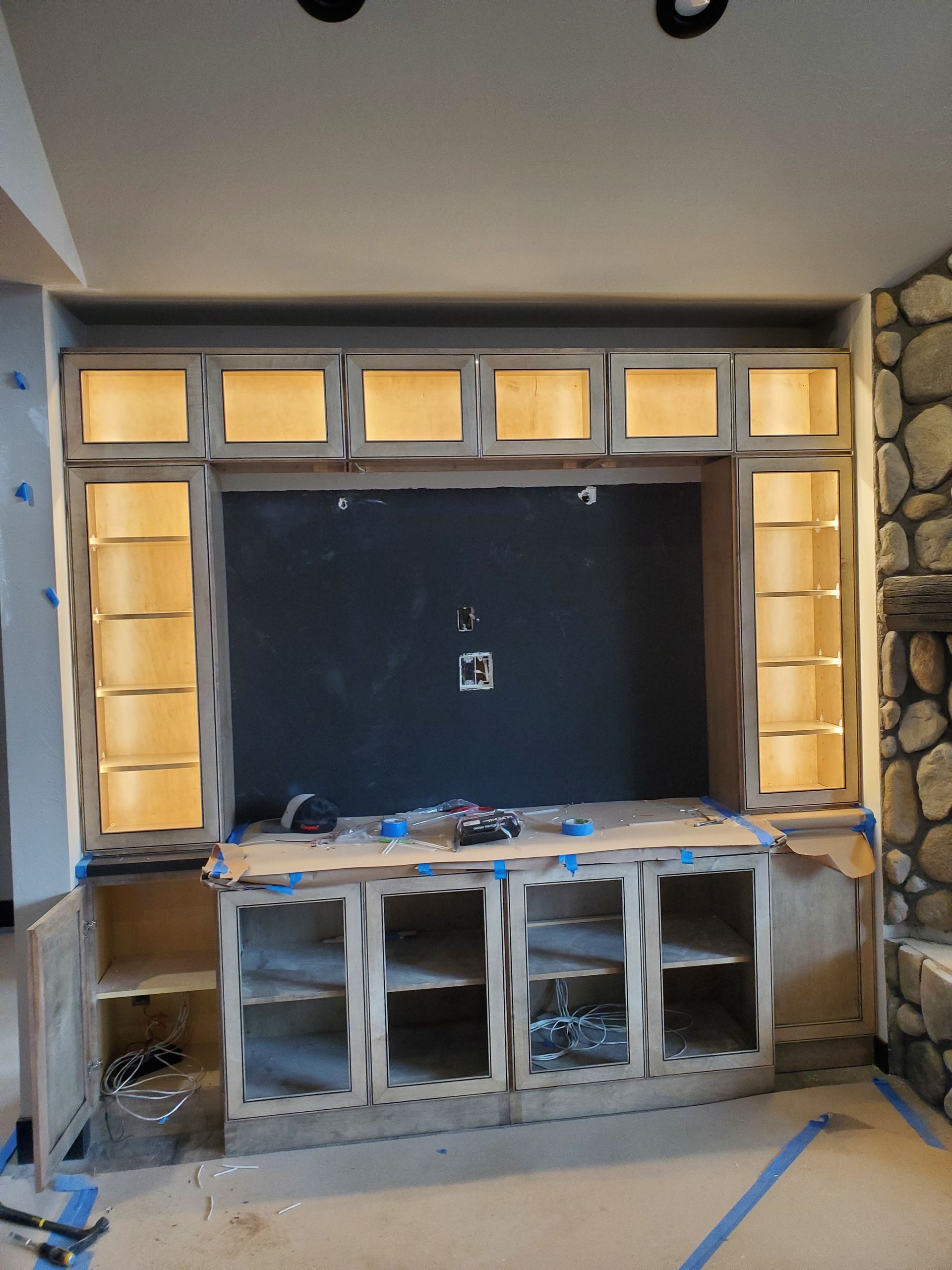 Wooden built-in entertainment center, under construction. Cabinets and shelving are unfinished with open doors and a dark-painted TV space.