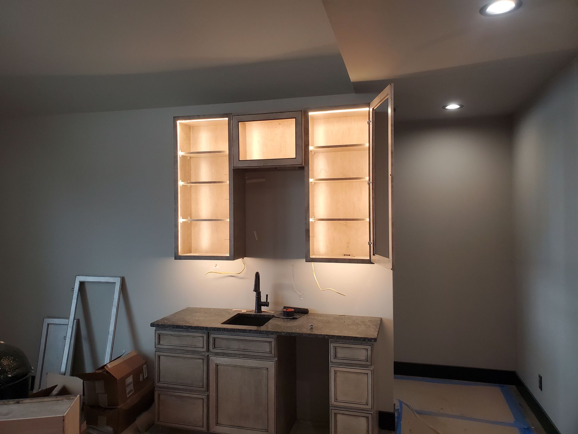 Built-in bar with upper cabinets lit, dark sink, countertop, and lower cabinets. A construction site, interior.