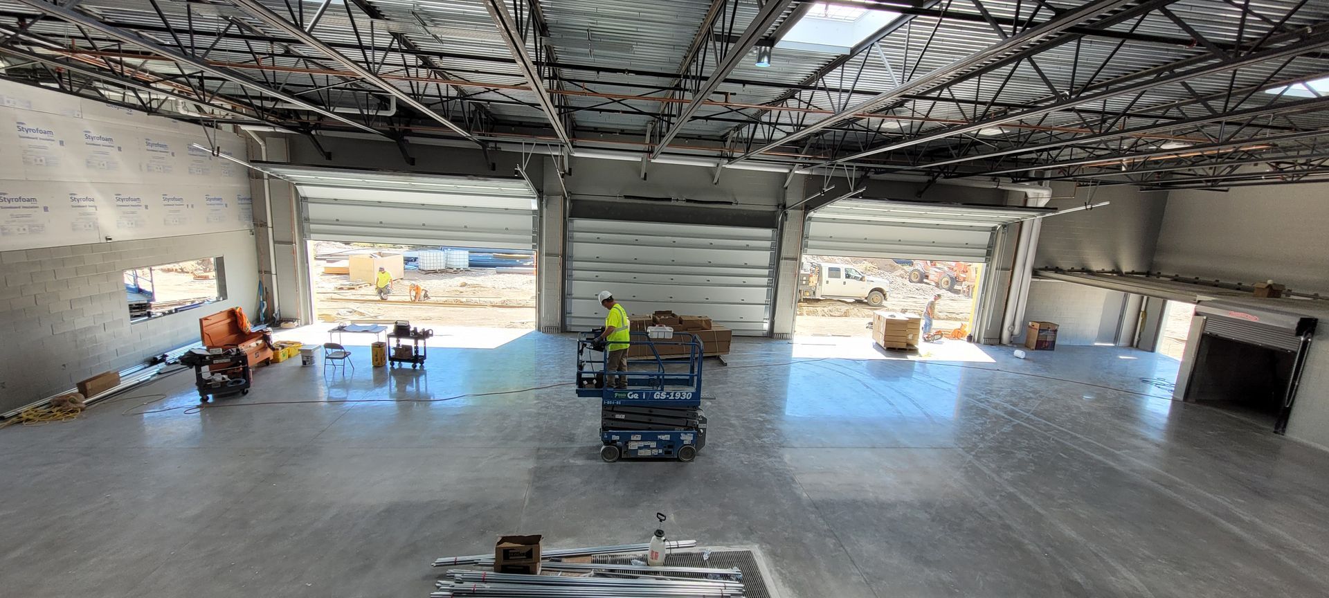 Construction site interior with open garage doors. A worker in a yellow vest operates equipment on the shiny concrete floor.