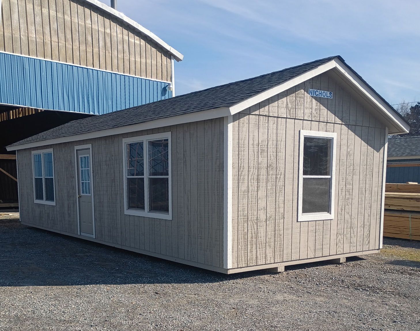 Gray cabin with black roof and white-framed windows, set on gravel. Another building in background.