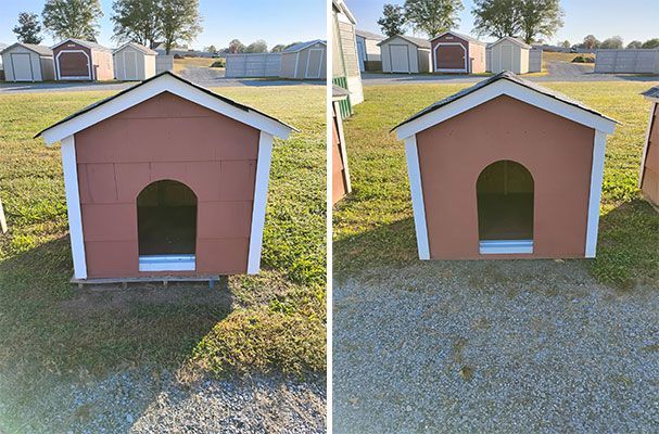 Two brown dog houses with white trim, sitting on grass and gravel, with a row of sheds in the background.