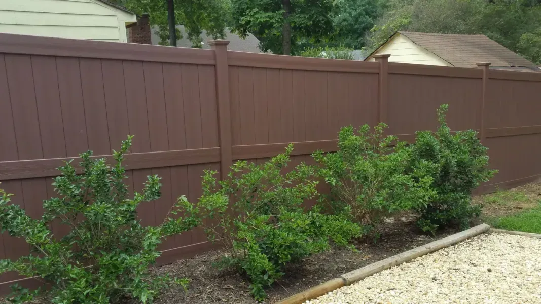 A brown wooden fence is surrounded by bushes and gravel in a backyard.