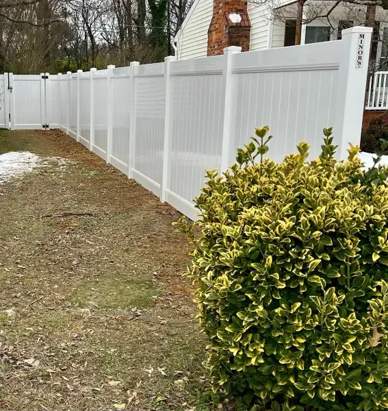 A white fence with a bush in the foreground and a house in the background.