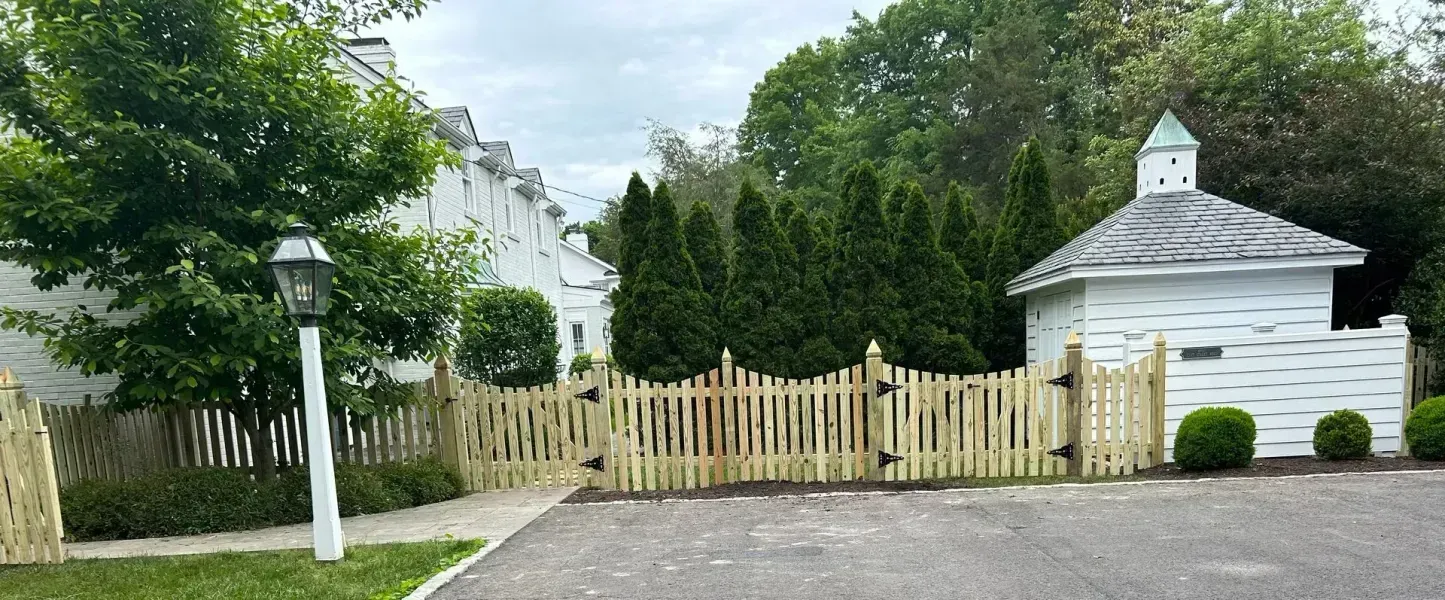 A wooden fence surrounds a driveway leading to a white house.
