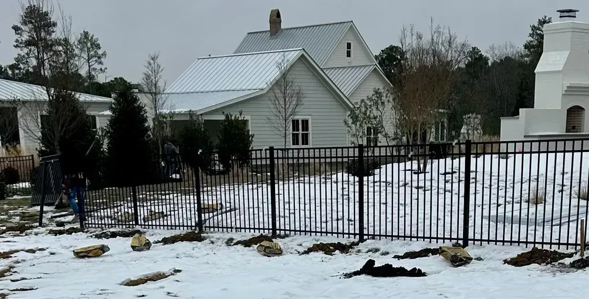A snowy yard with a black fence and a white house in the background.