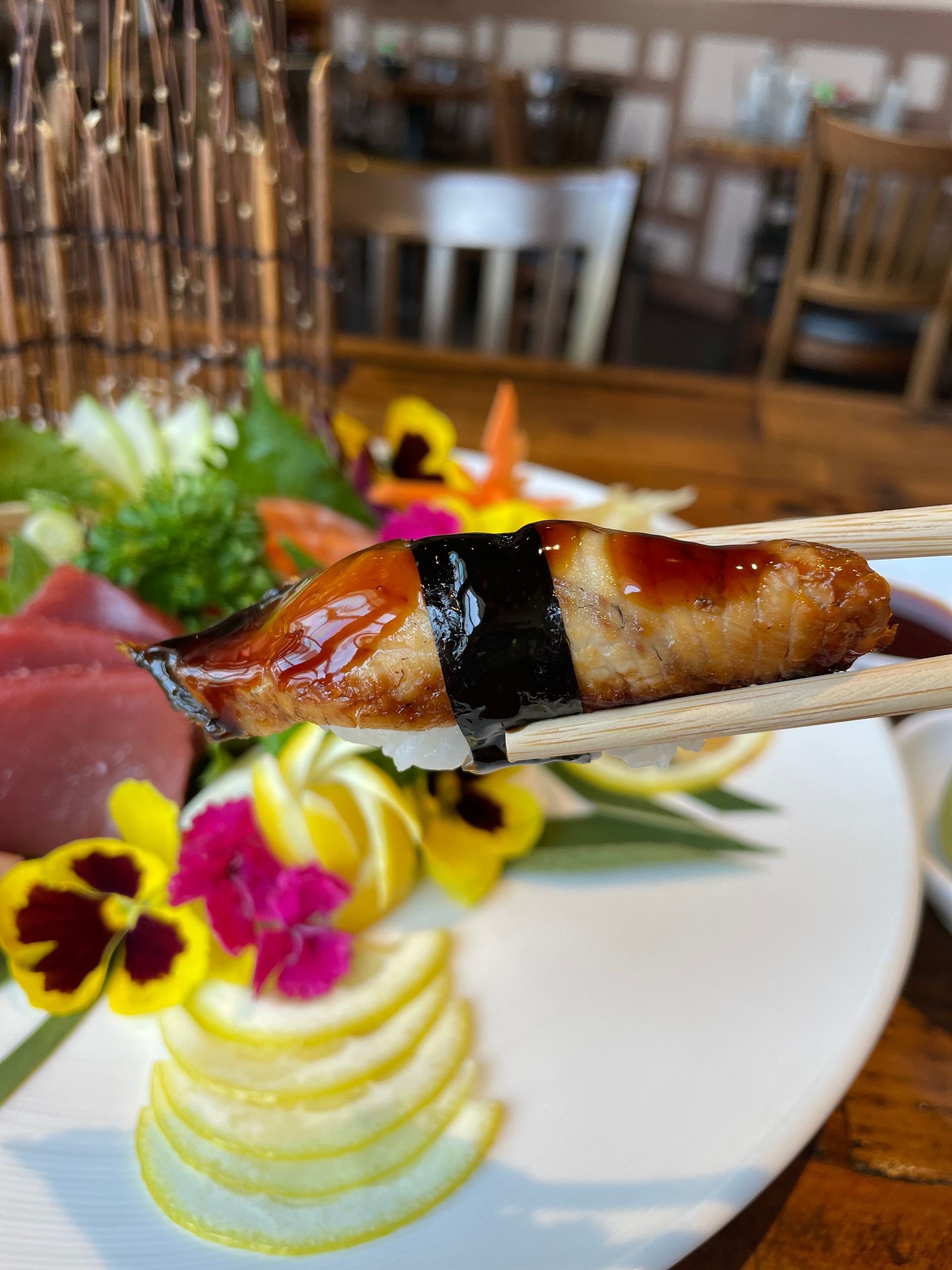 A person is holding a piece of sushi with chopsticks over a plate of food
