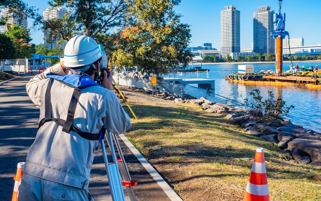 A surveyor in a hard hat and high-visibility vest uses a transit level by a river near city buildings and a construction site.