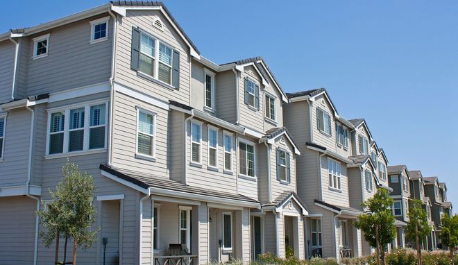 A row of multi-story, beige suburban townhouses with gabled roofs and windows under a clear blue sky.