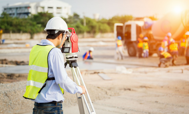 A construction surveyor in a white hard hat and yellow vest uses a tripod-mounted total station at a sunny job site.