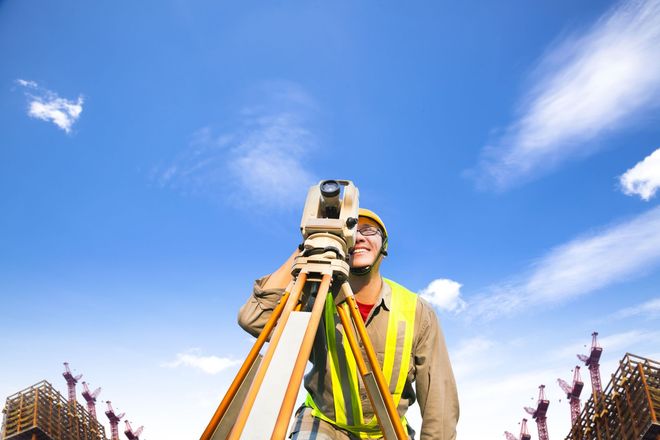 A construction surveyor in a yellow safety vest looks through a theodolite on a tripod against a bright blue sky.
