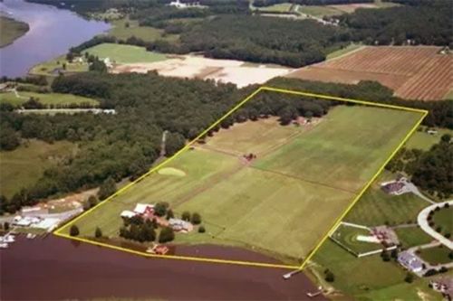 Aerial view of a rectangular property outlined in yellow, featuring a house by a river, open fields, and surrounding trees.