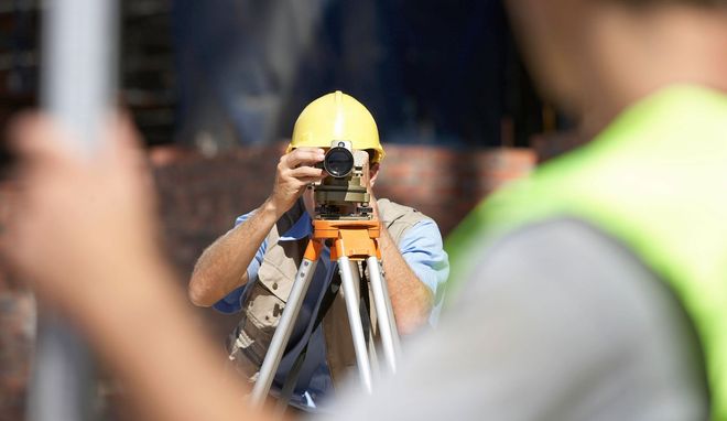 A person in a hard hat looks through a surveying instrument mounted on a tripod, with another worker in the foreground.