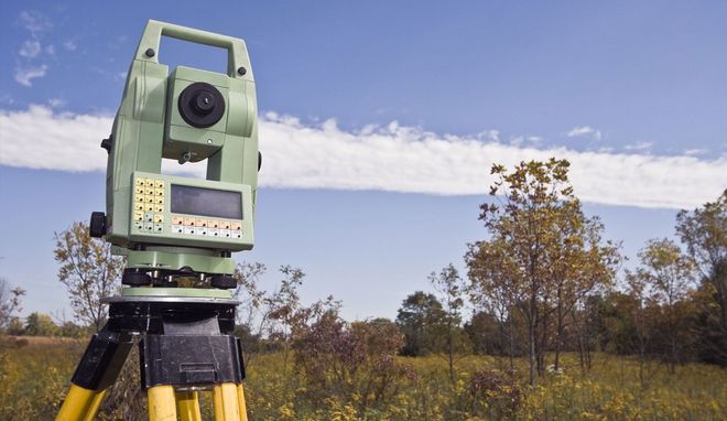A light green surveying total station mounted on a tripod in a grassy field under a blue sky with thin clouds.