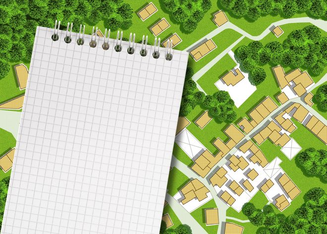 A large, blank spiral-bound notepad sits atop a top-down view of a small, green village with multiple buildings.