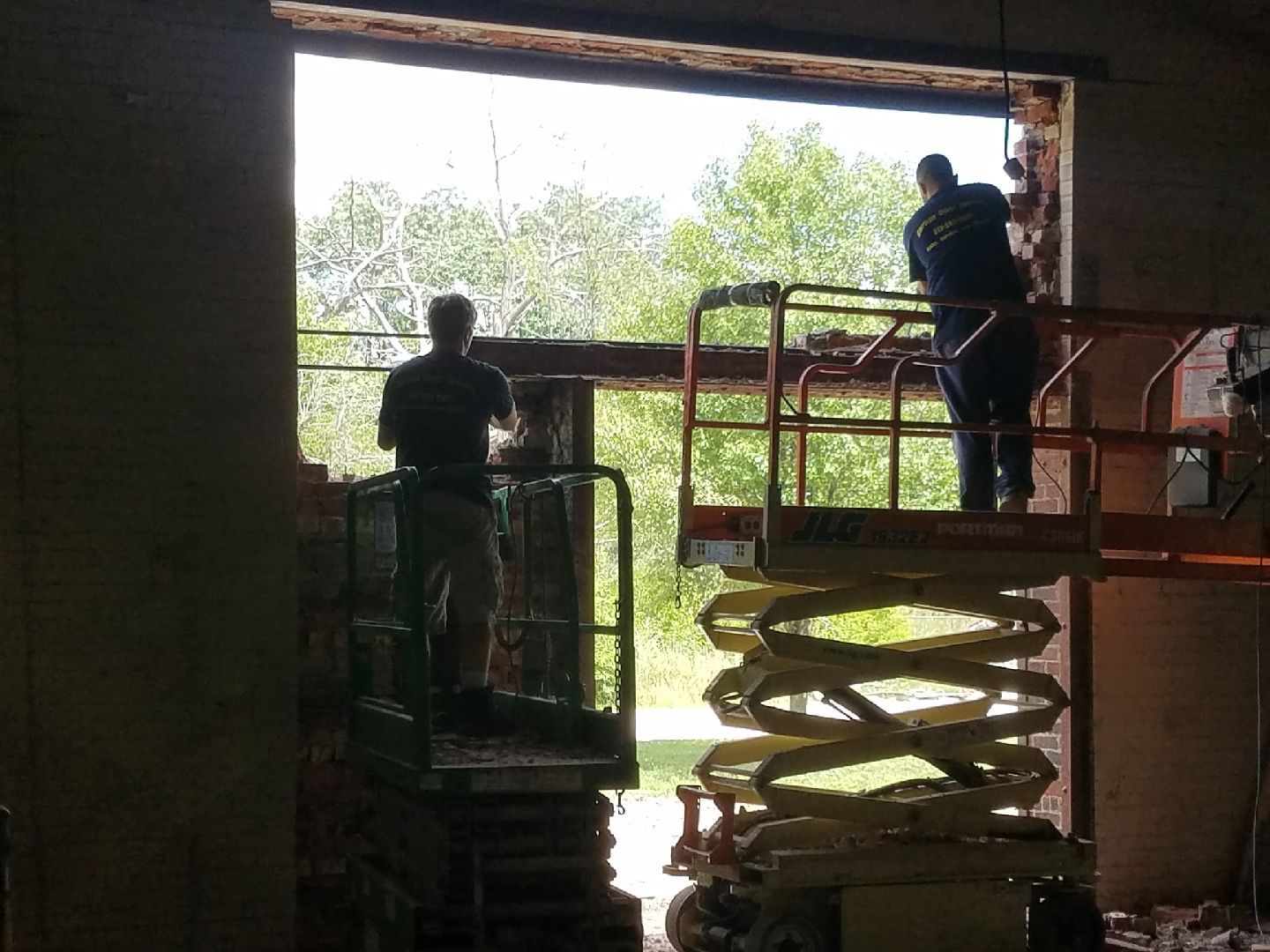 Two workers on scissor lifts installing a large window opening inside a building.