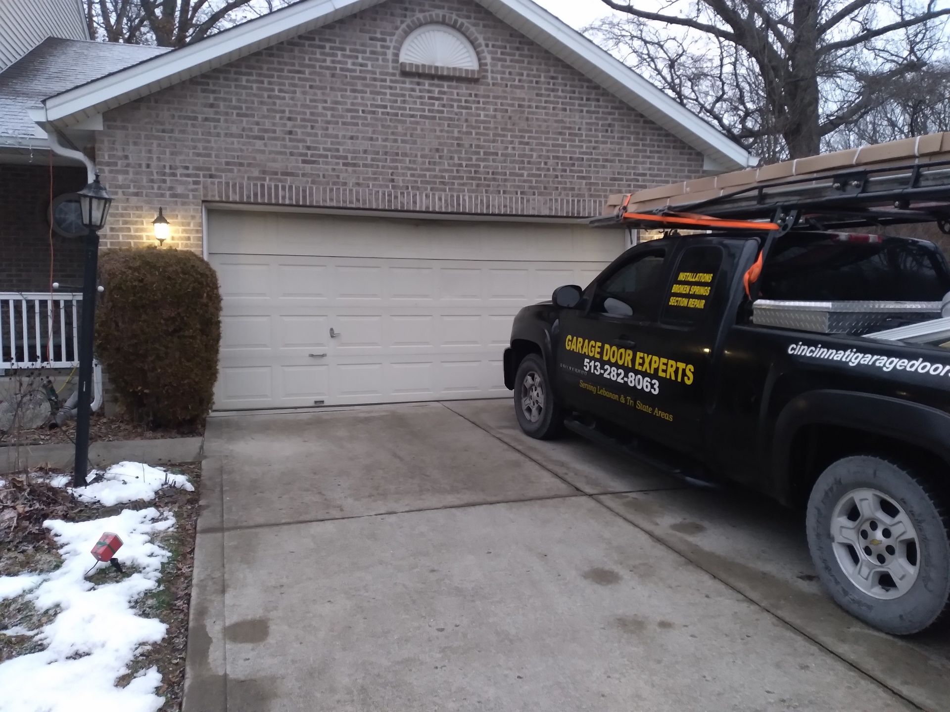 Black service truck parked in a driveway in front of a beige garage on a snowy suburban street