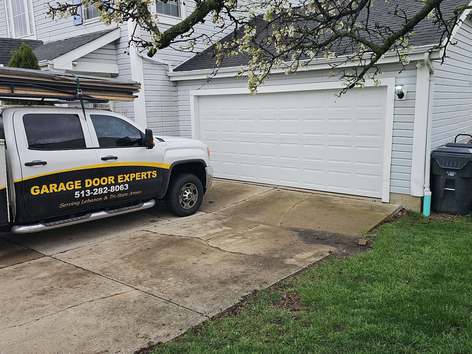 White garage door beside a white work truck parked in a driveway with wet concrete and a tree overhead