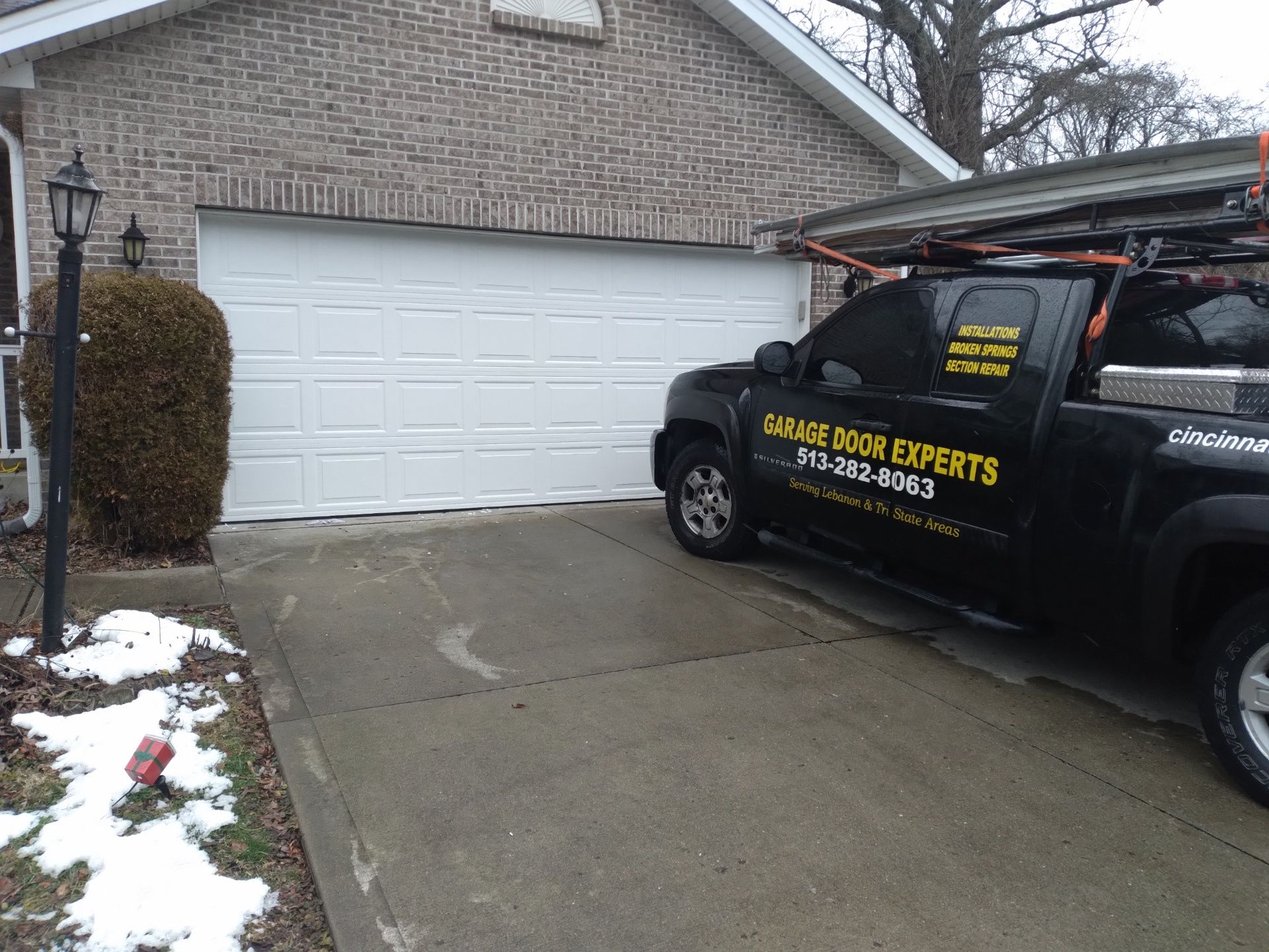 Black service truck parked in front of a garage beside a wet driveway with snow patches