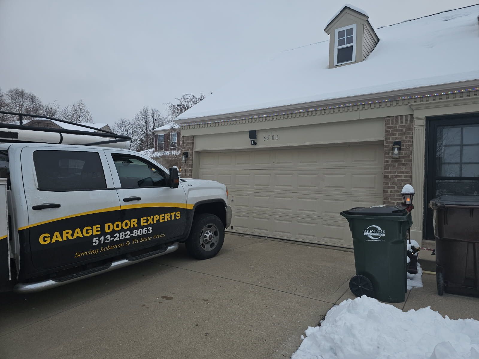 Garage door service van parked in a snowy driveway beside a beige house garage.