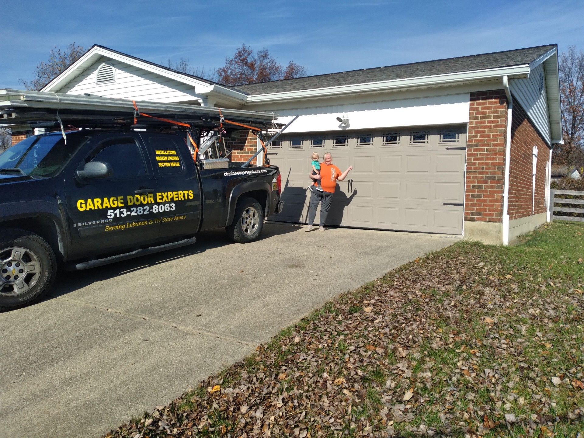 Garage door repair van parked in a driveway with two workers by a gray garage in a suburban home.