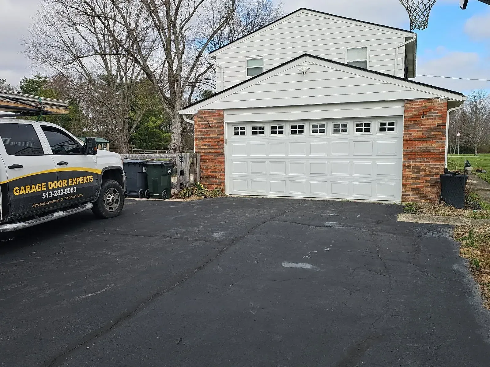 Suburban driveway with a white garage and a black-and-yellow utility truck parked beside trash bins.