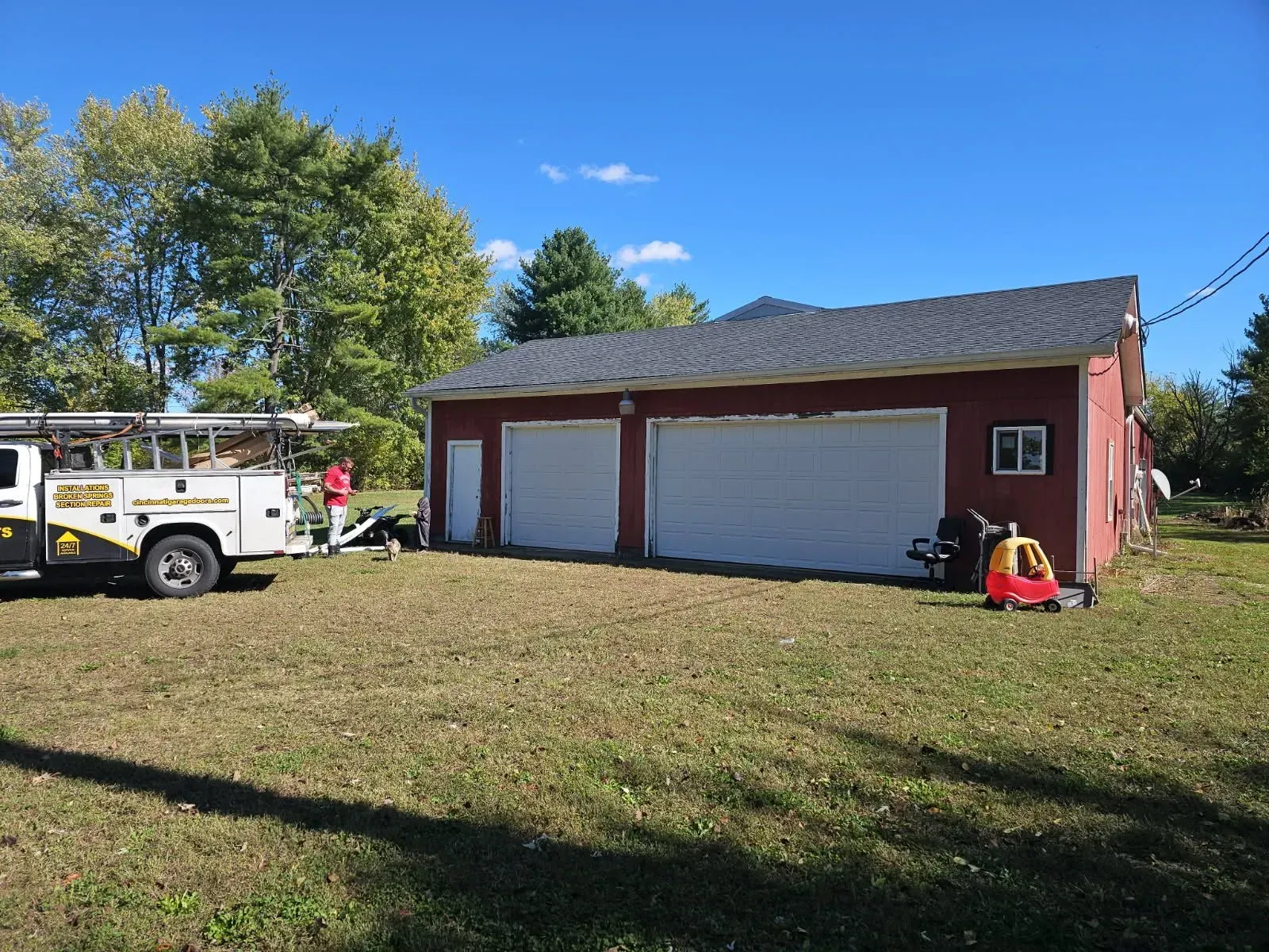 Red garage with white doors on a grassy lot, a utility truck parked beside it under a blue sky.