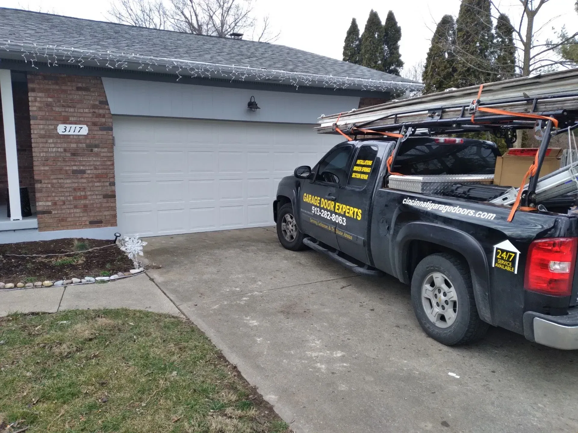 Black pickup with ladder rack parked in driveway beside a garage on a cloudy day