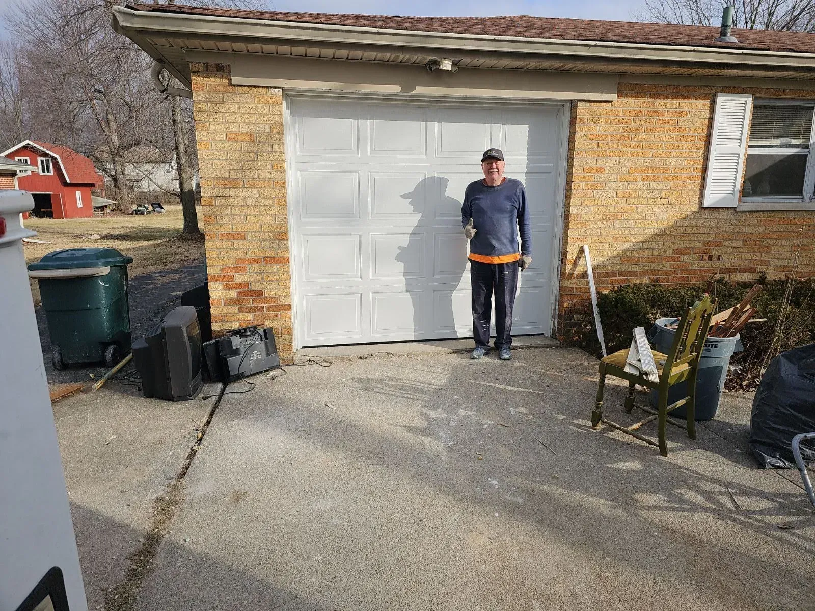Person standing in front of a white garage door beside a driveway with trash bins and folding chairs