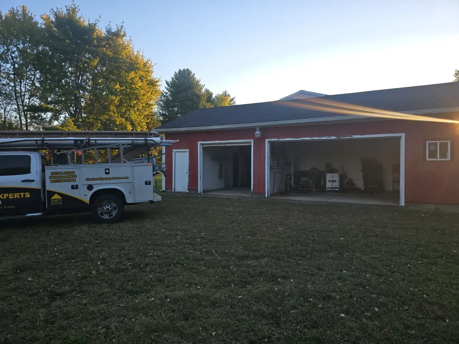 Fire truck parked beside a red garage with open bays in a grassy yard at sunset.