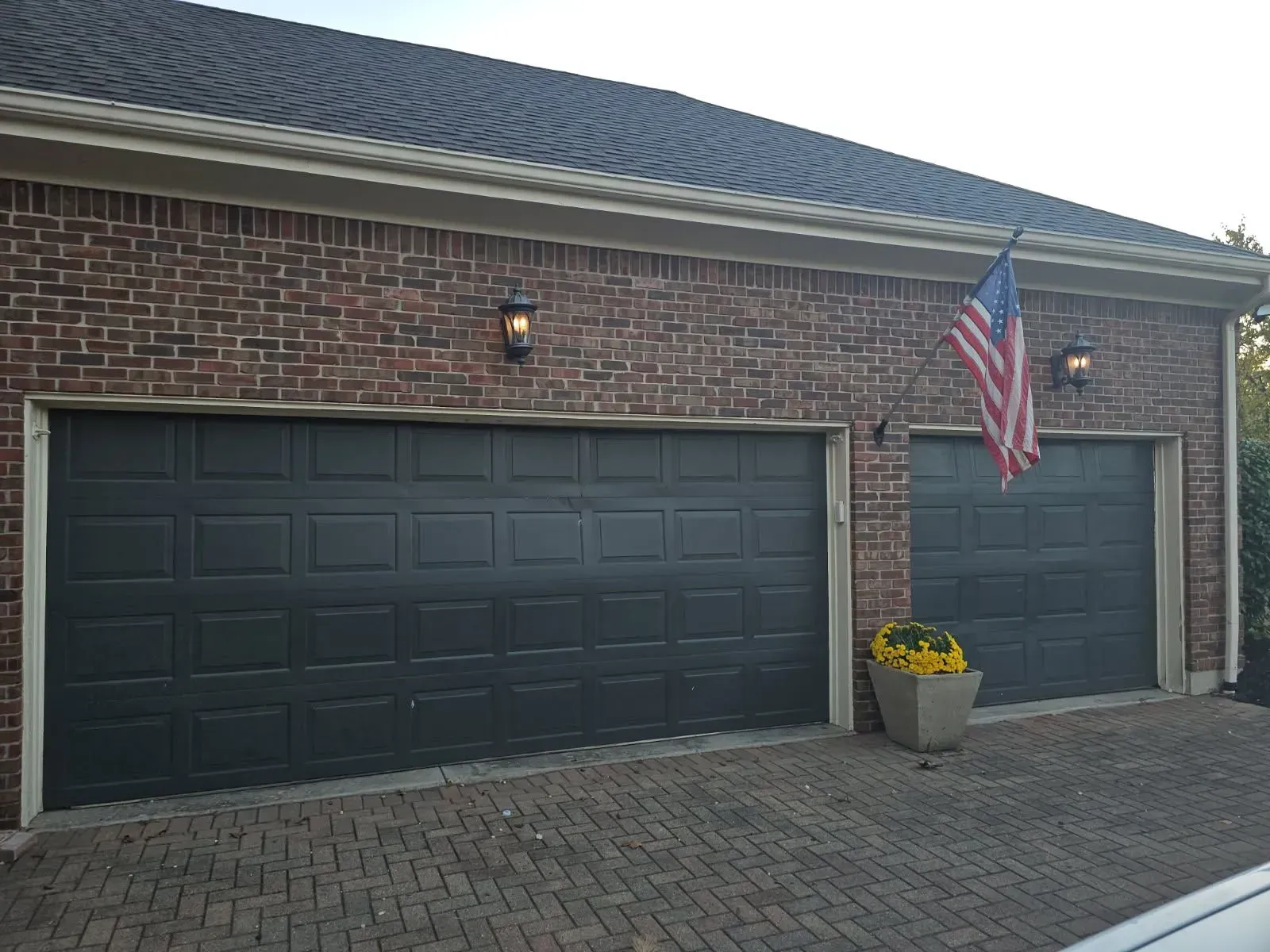 Brick house front with black garage doors, a U.S. flag, and potted flowers by the driveway