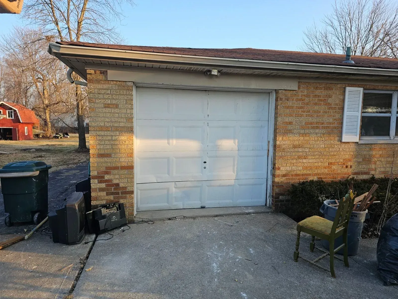 Small brick garage with a white door beside a driveway, bins, and a chair outside.