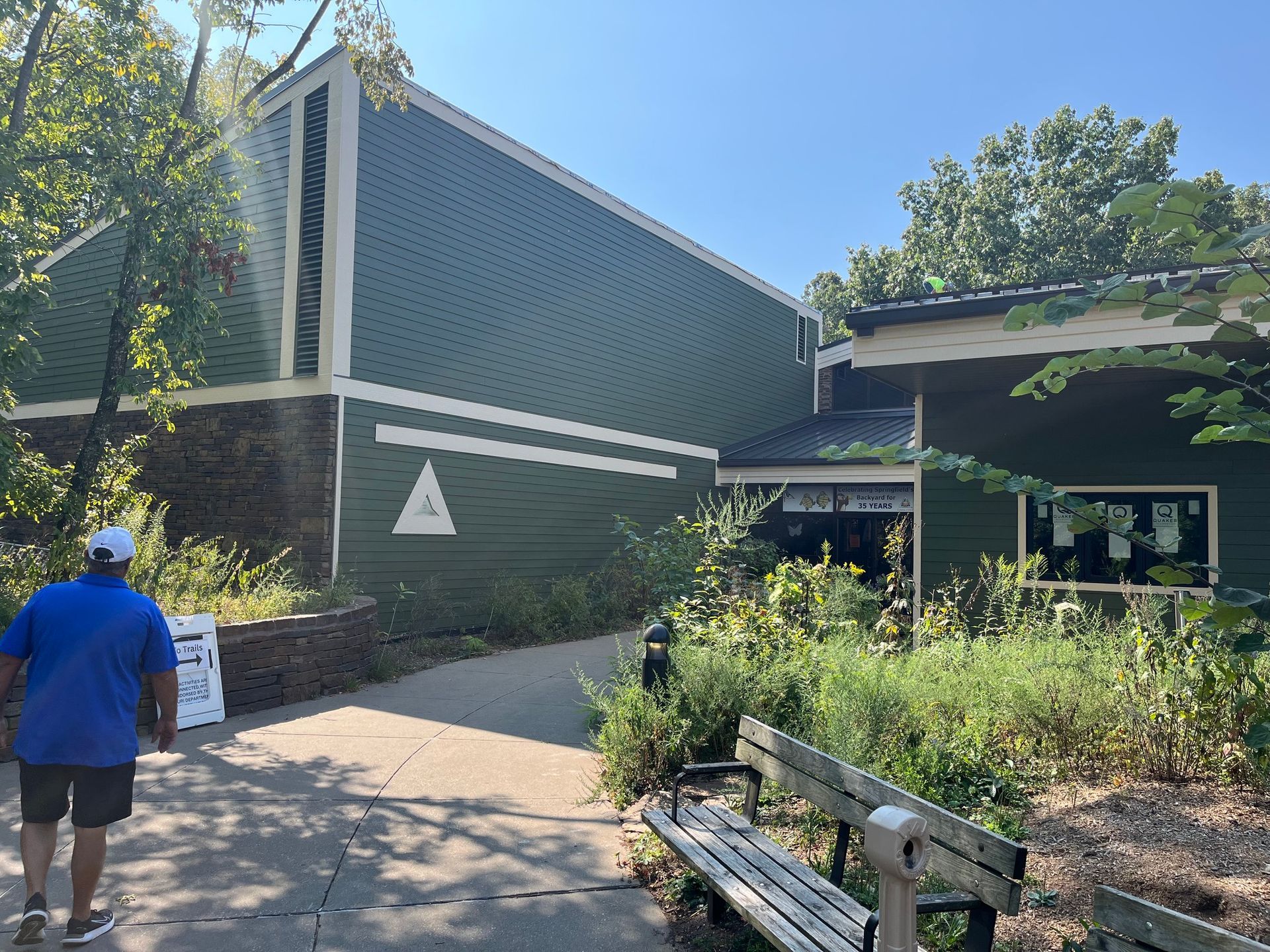 Green building with triangular logo and path leading to entrance. Man walks towards building.