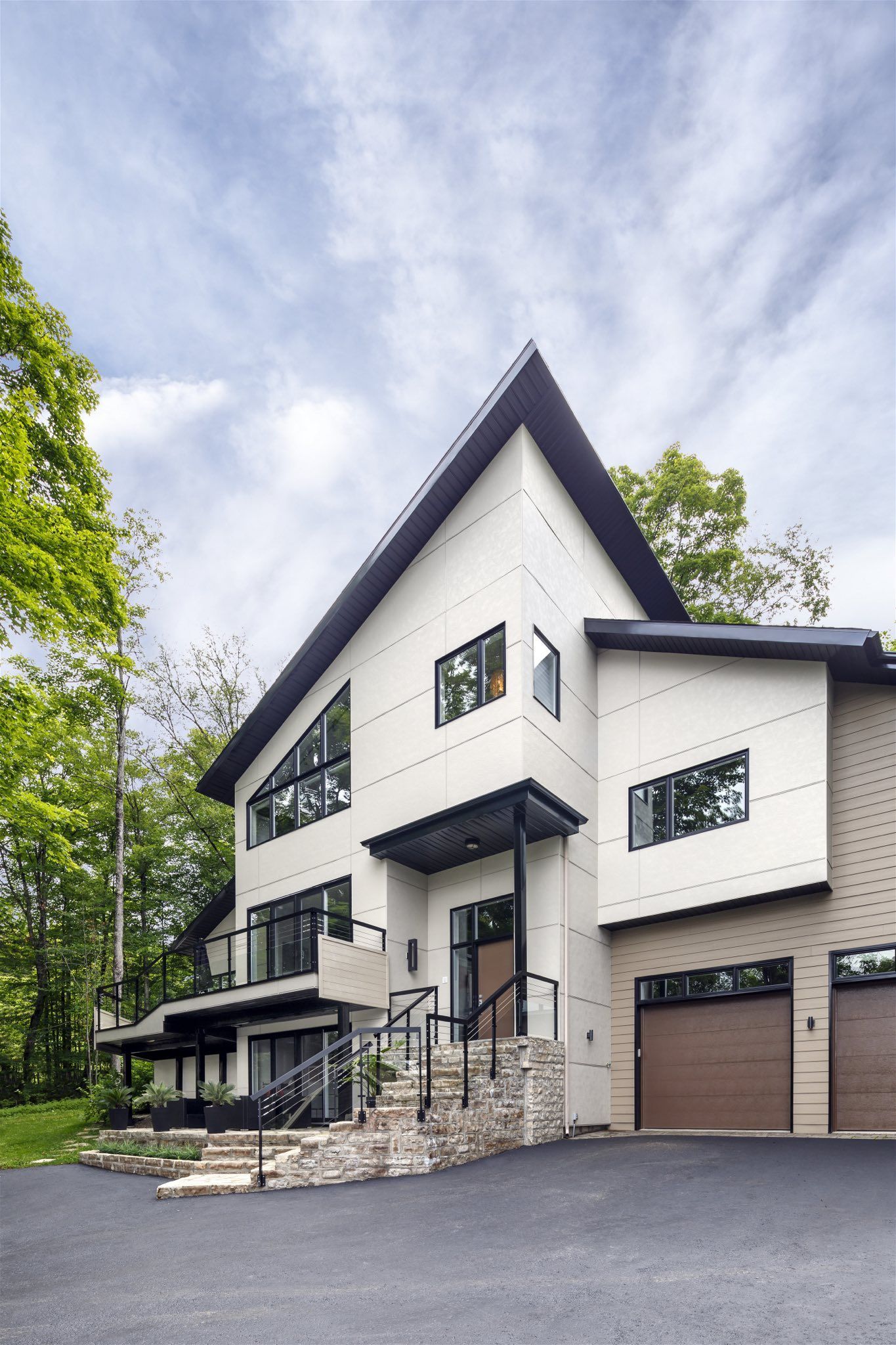 Modern white house with black accents, a sloped roof, and a two-car garage. Set in a wooded area.