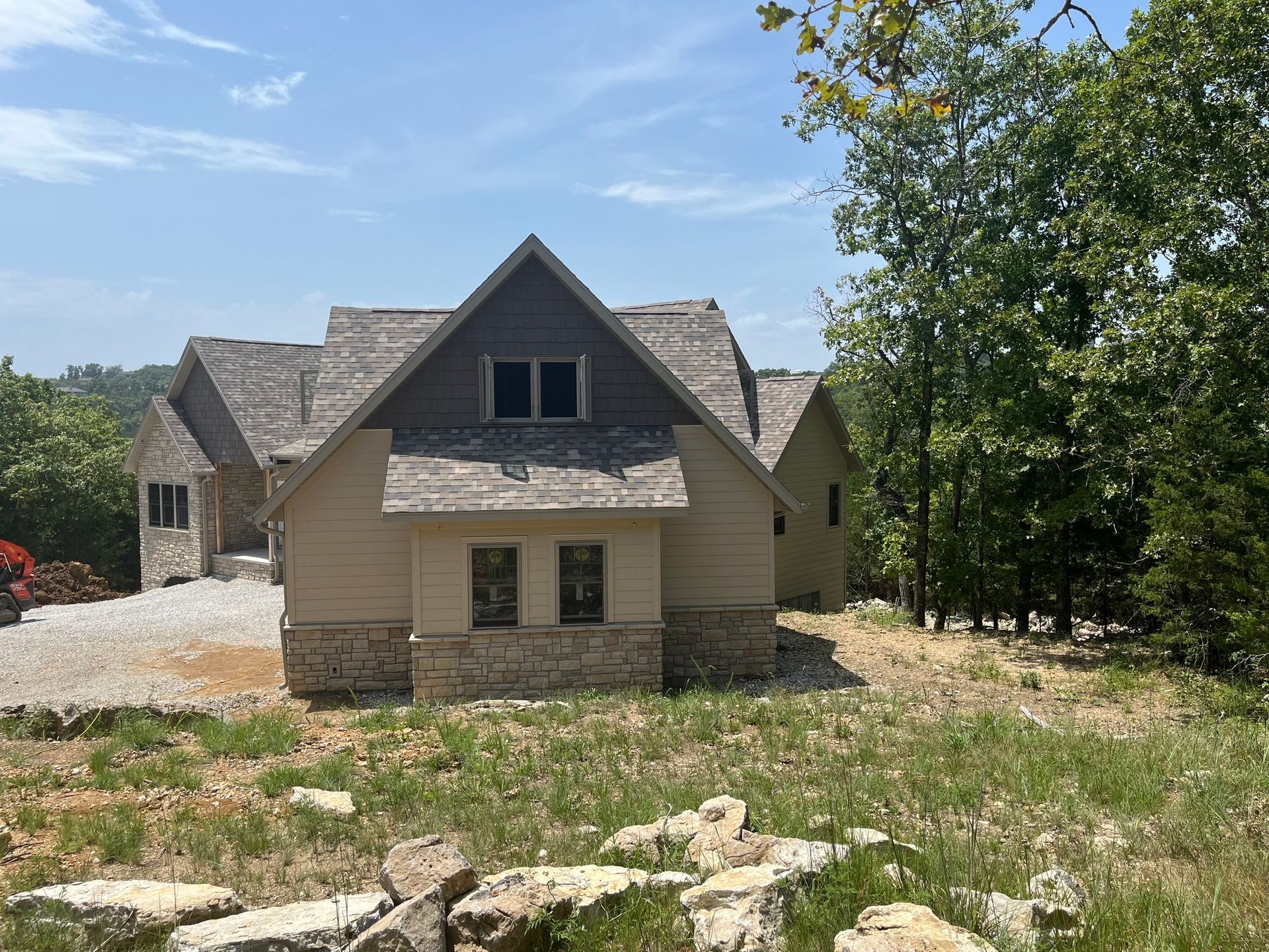 Beige and stone house with shingled roof, set on a grassy hill surrounded by trees under a blue sky.