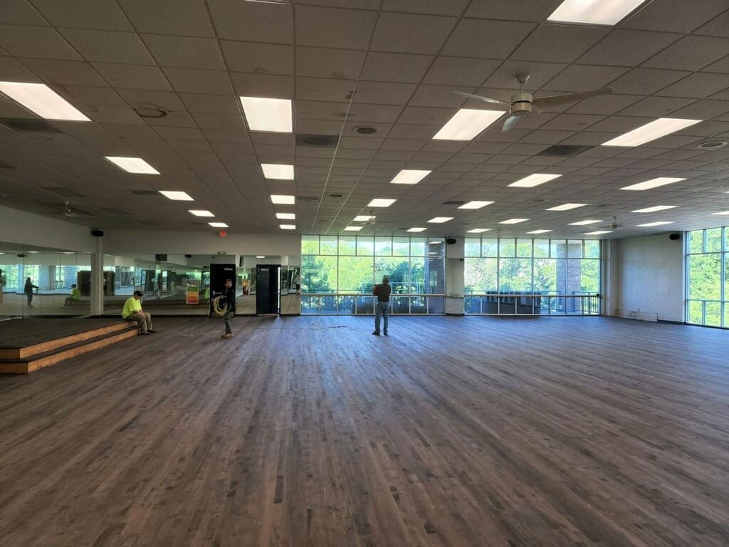 Large empty studio with wood floors, large windows, and overhead lights; a few people are present.