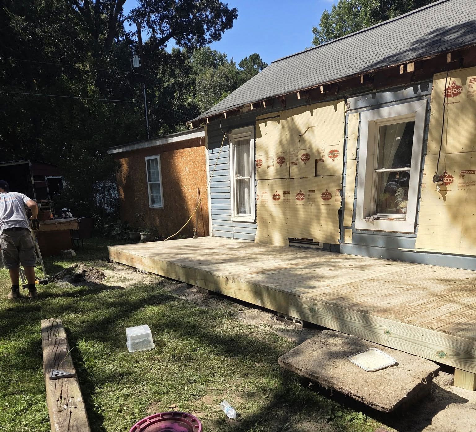 Construction of a wooden deck next to a house with exposed siding and a worker.