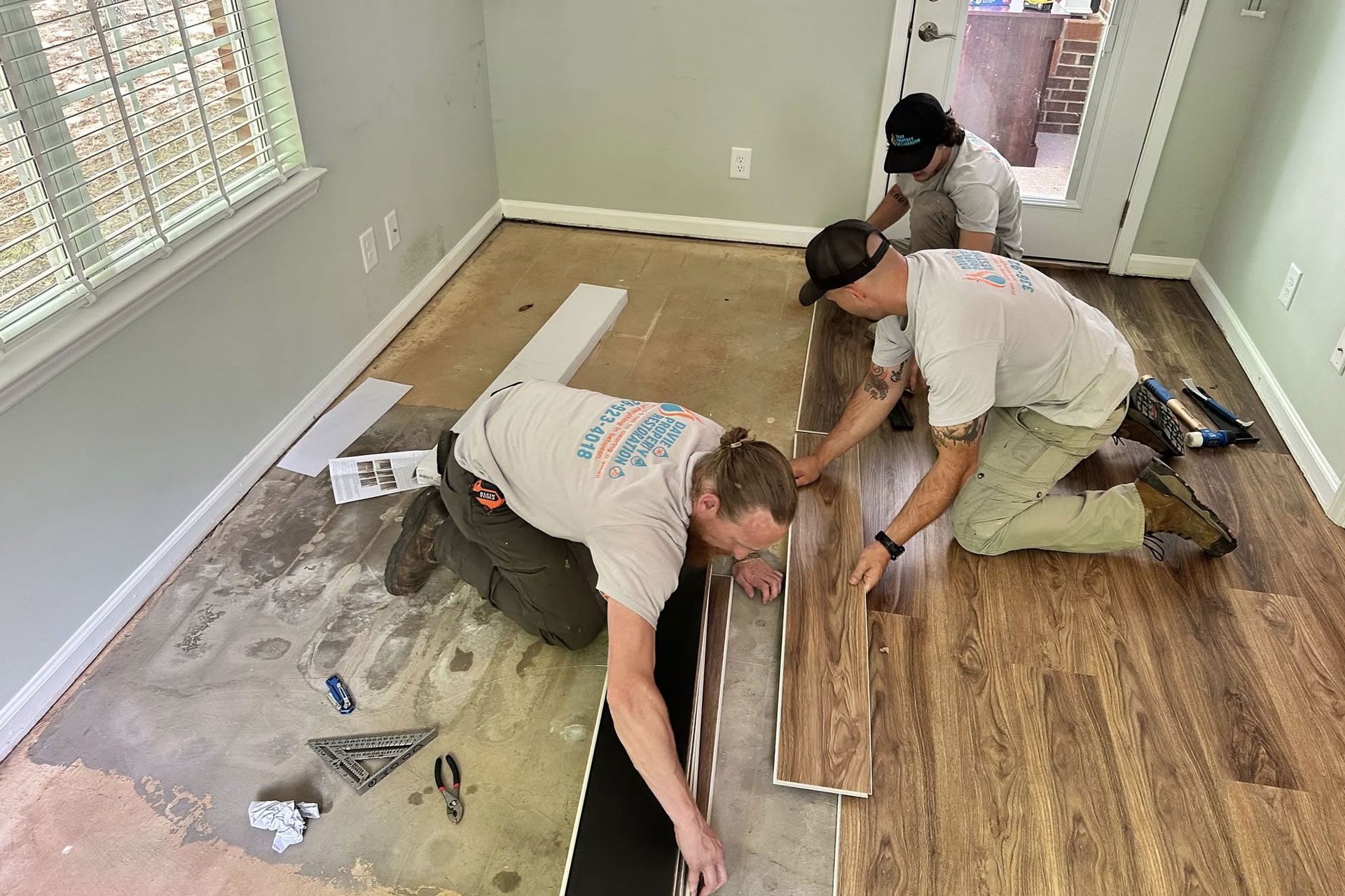 Three workers installing wood-look flooring in a room with a window and doorway.