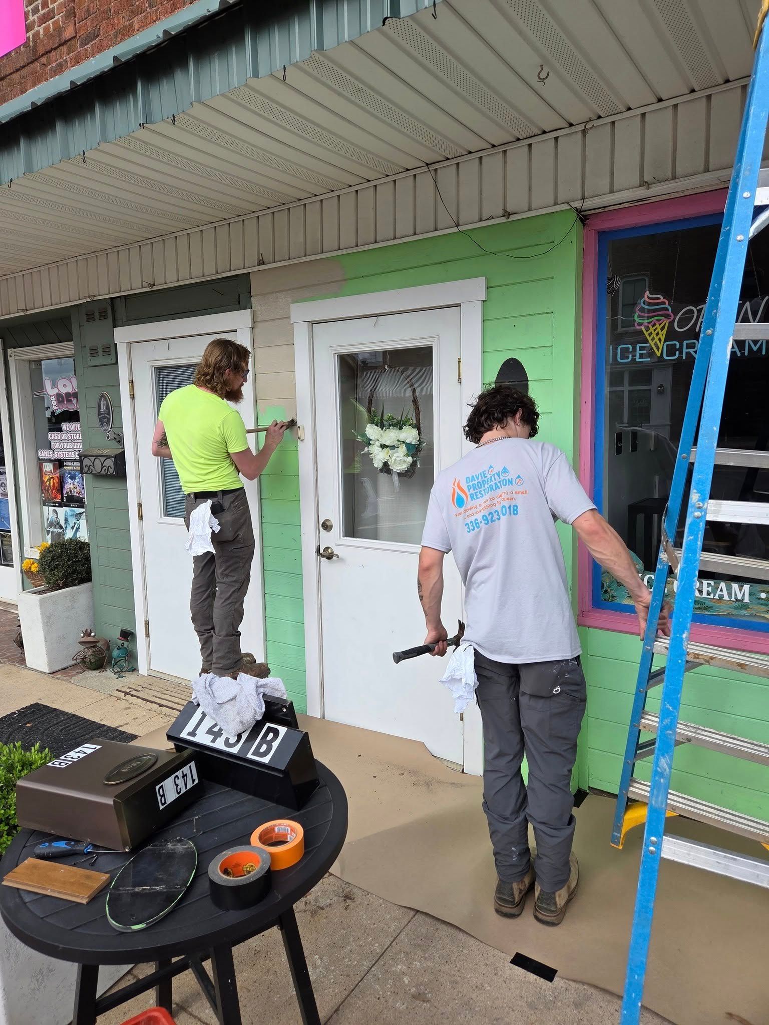 Two people painting a storefront; one on a ladder, one standing on a table.