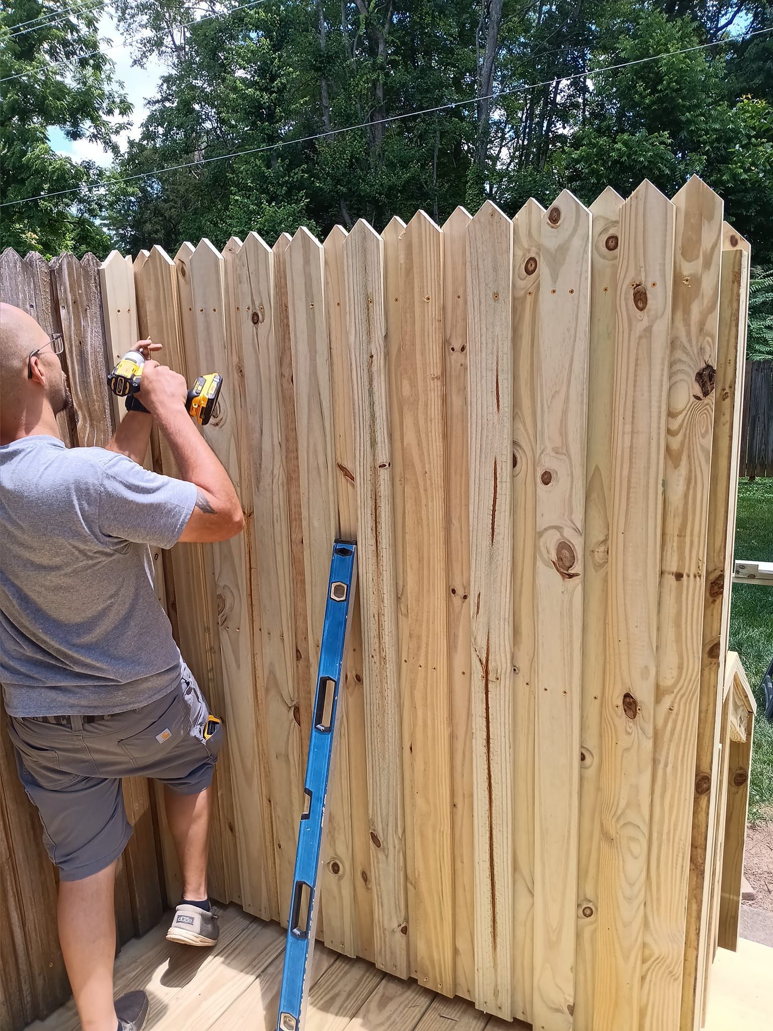 Person using a drill to attach a wooden picket fence. A level rests against the fence.