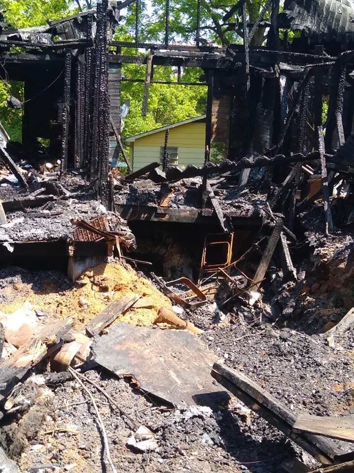 Charred remains of a building after a fire, showing burnt wood and rubble. A yellow structure is visible in the background.