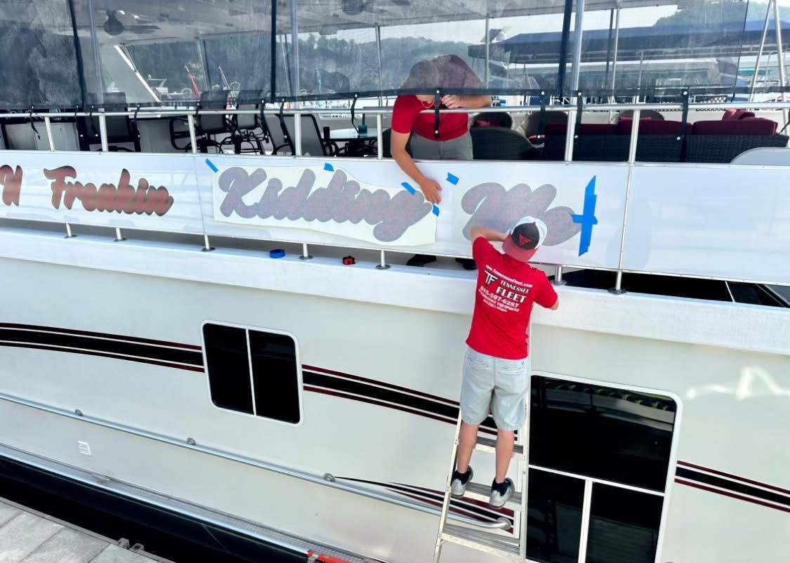 A man in a red shirt is painting a sign on the side of a boat