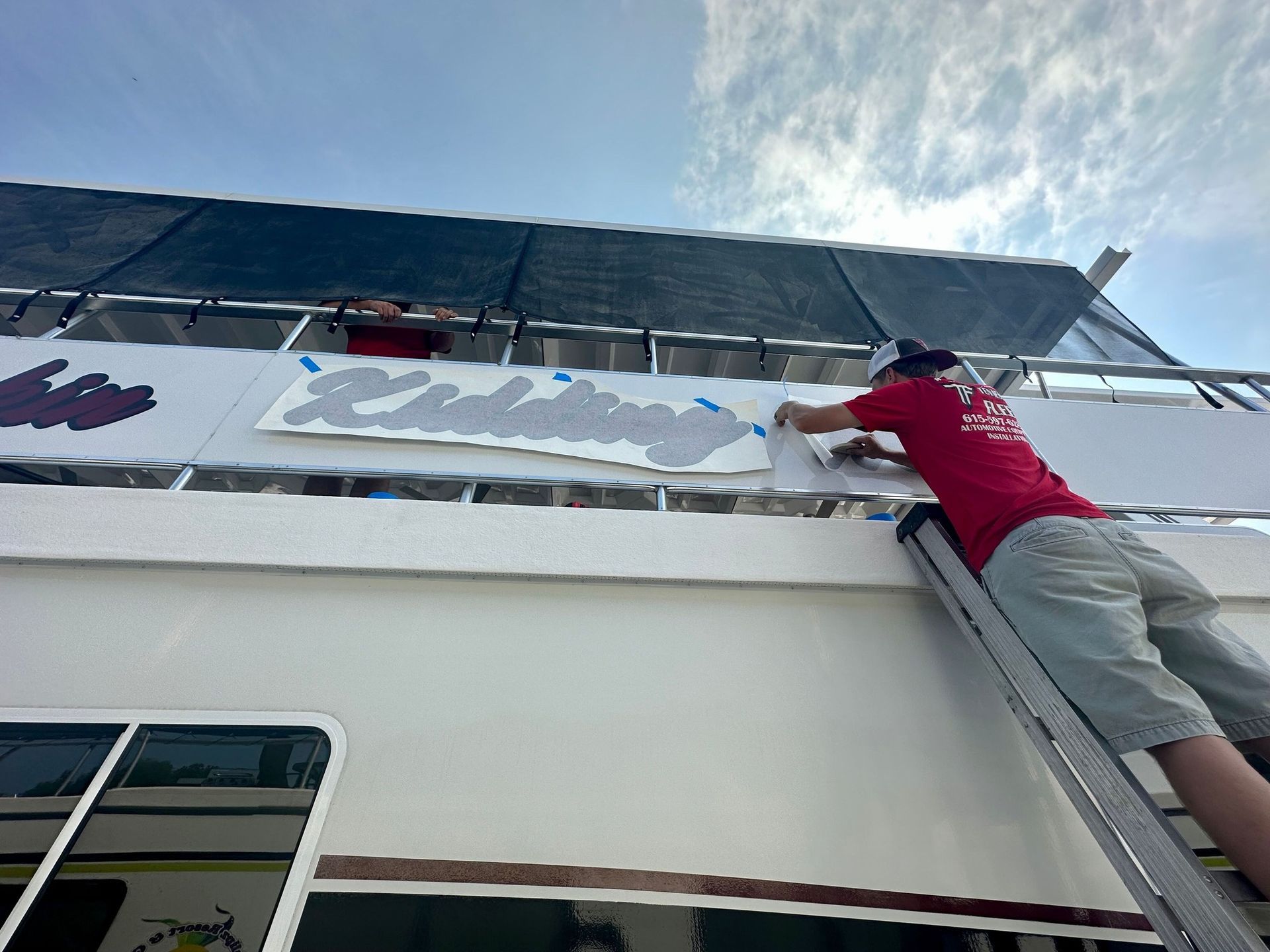 A man is standing on a ladder painting a sign on the side of a boat.