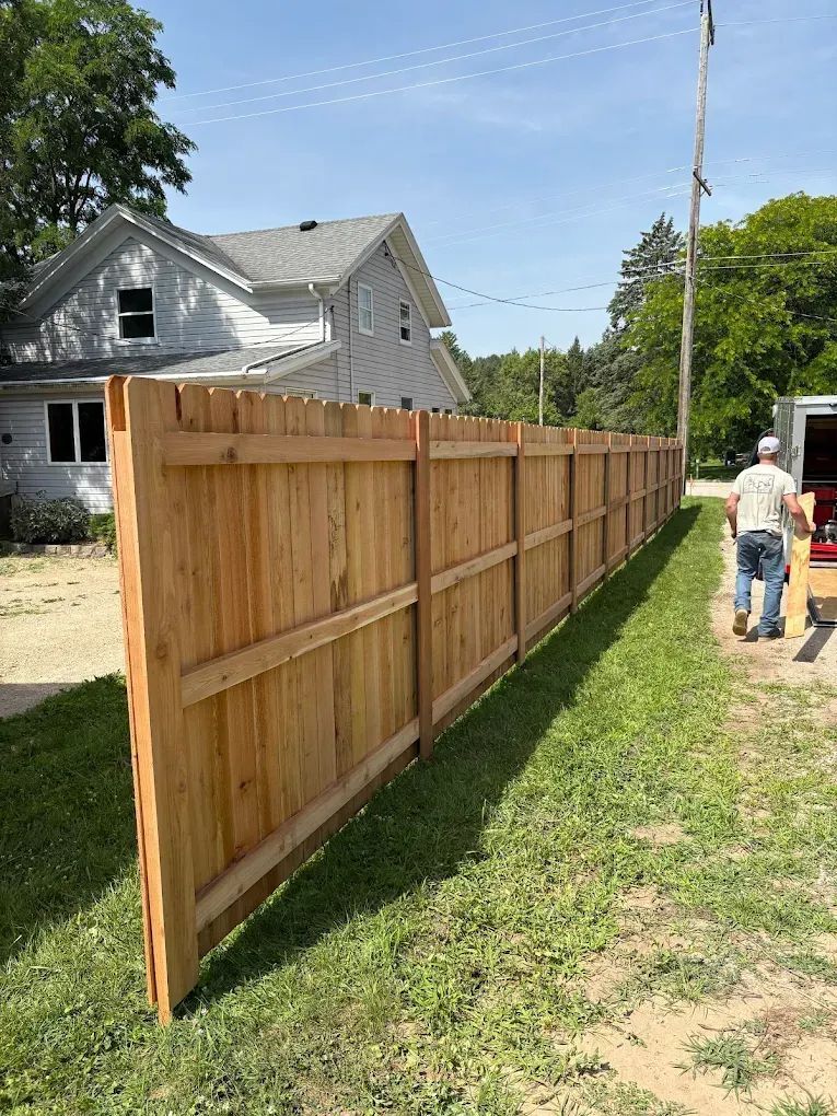 A long wooden fence in front of a house, with a person carrying items alongside. Sunny day.