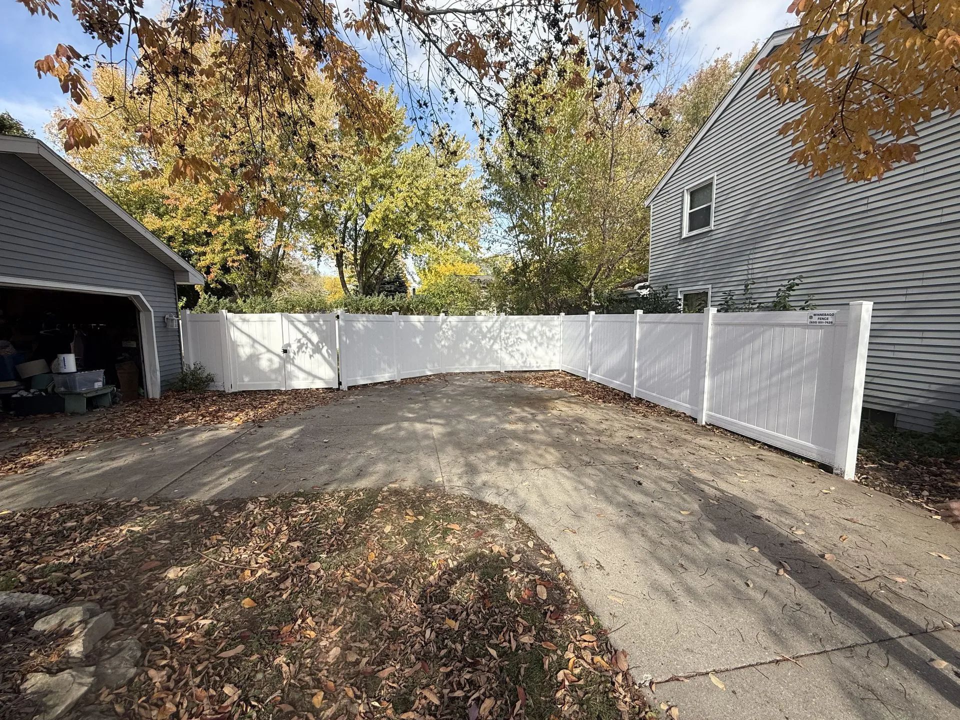White vinyl fence surrounding a driveway, next to a garage and house. Fall foliage overhead.