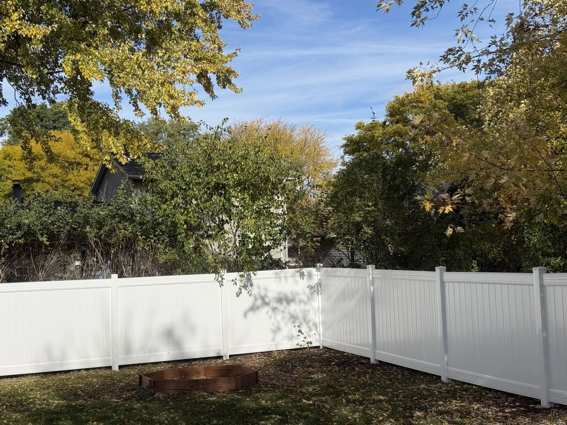 White fence surrounds a grassy yard with autumn trees and a partly cloudy sky.