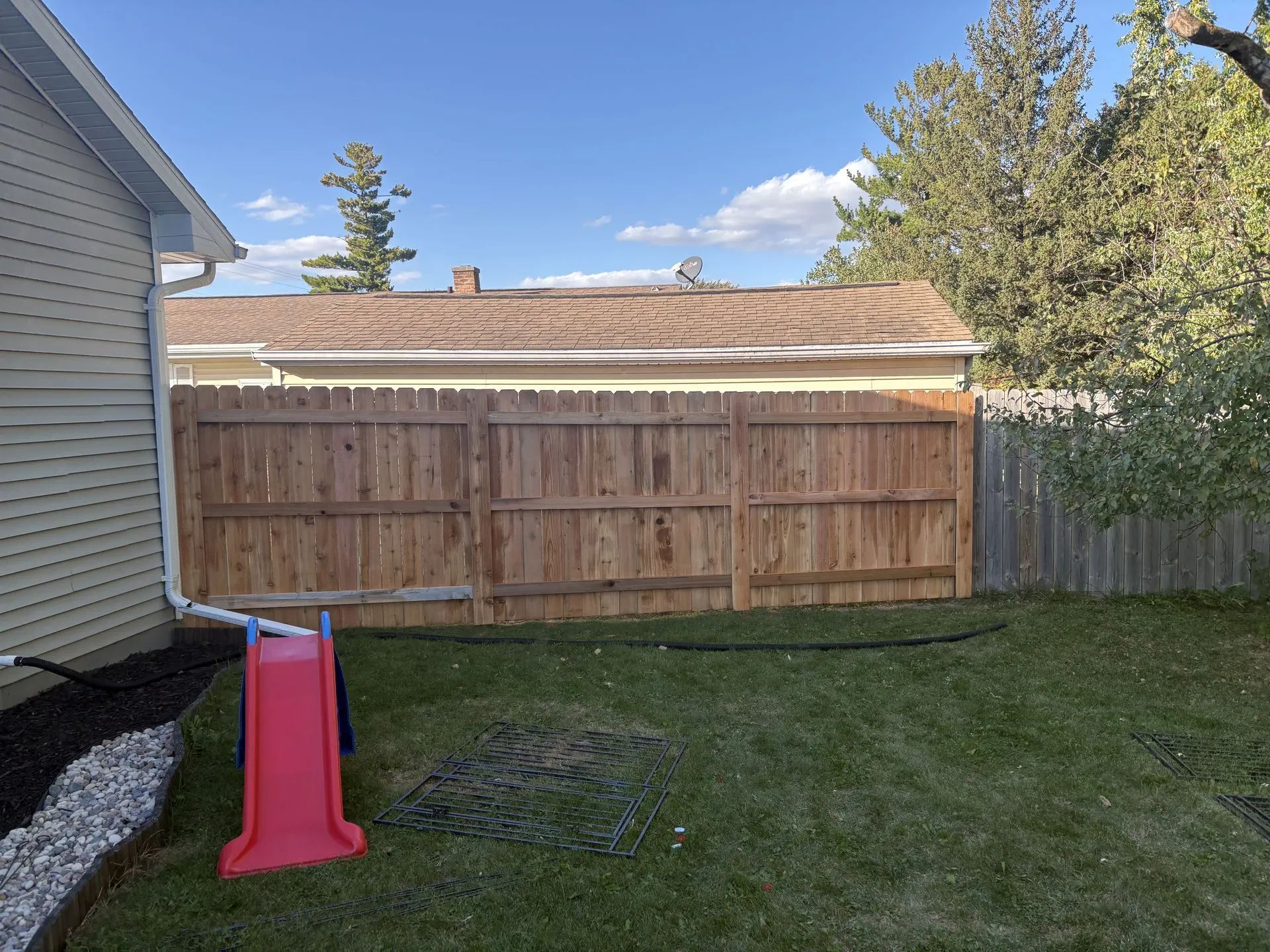 Backyard with a brown wooden fence, lawn, a red slide, and a glimpse of a house with a brown roof.