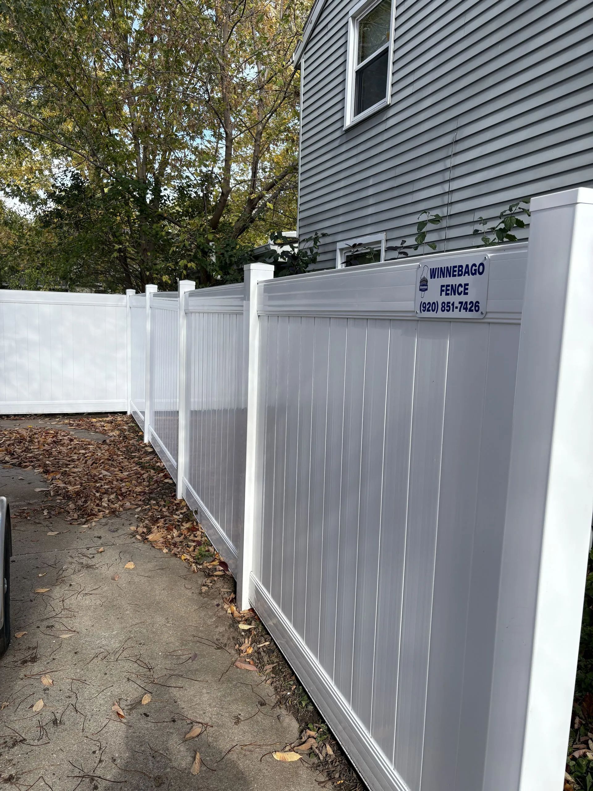 White vinyl fence along a driveway next to a gray house. Fall leaves are on the ground.
