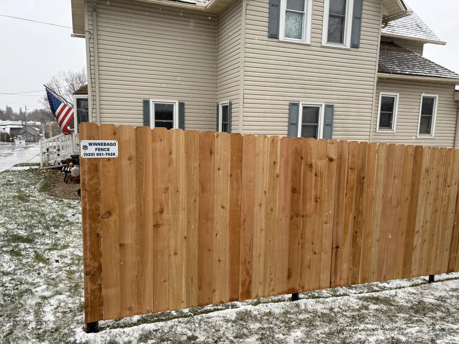 Wooden fence in front of a beige house with an American flag; snow on the ground.