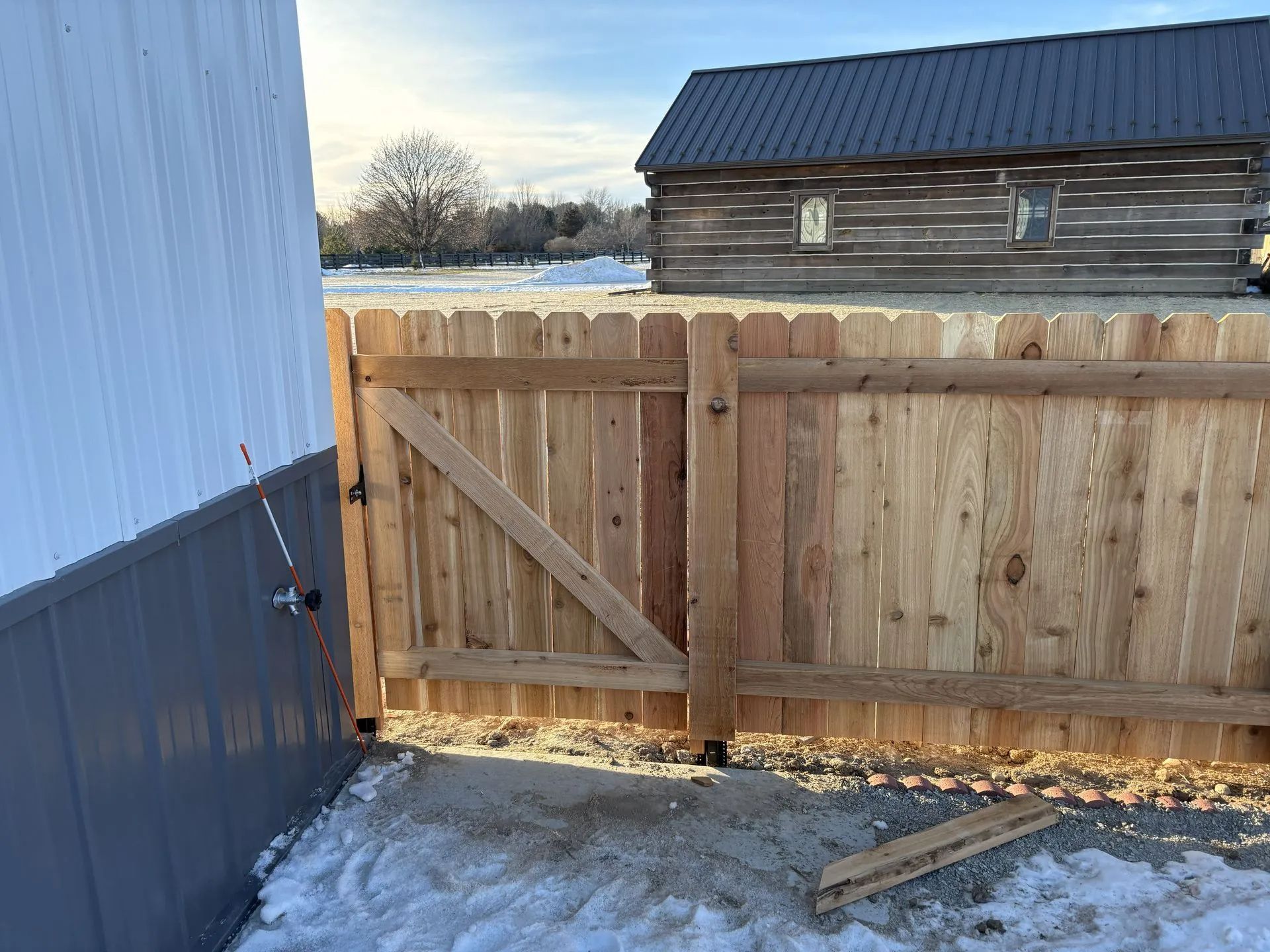 Wooden fence gate next to a building, in front of a log cabin on a sunny day with snow.
