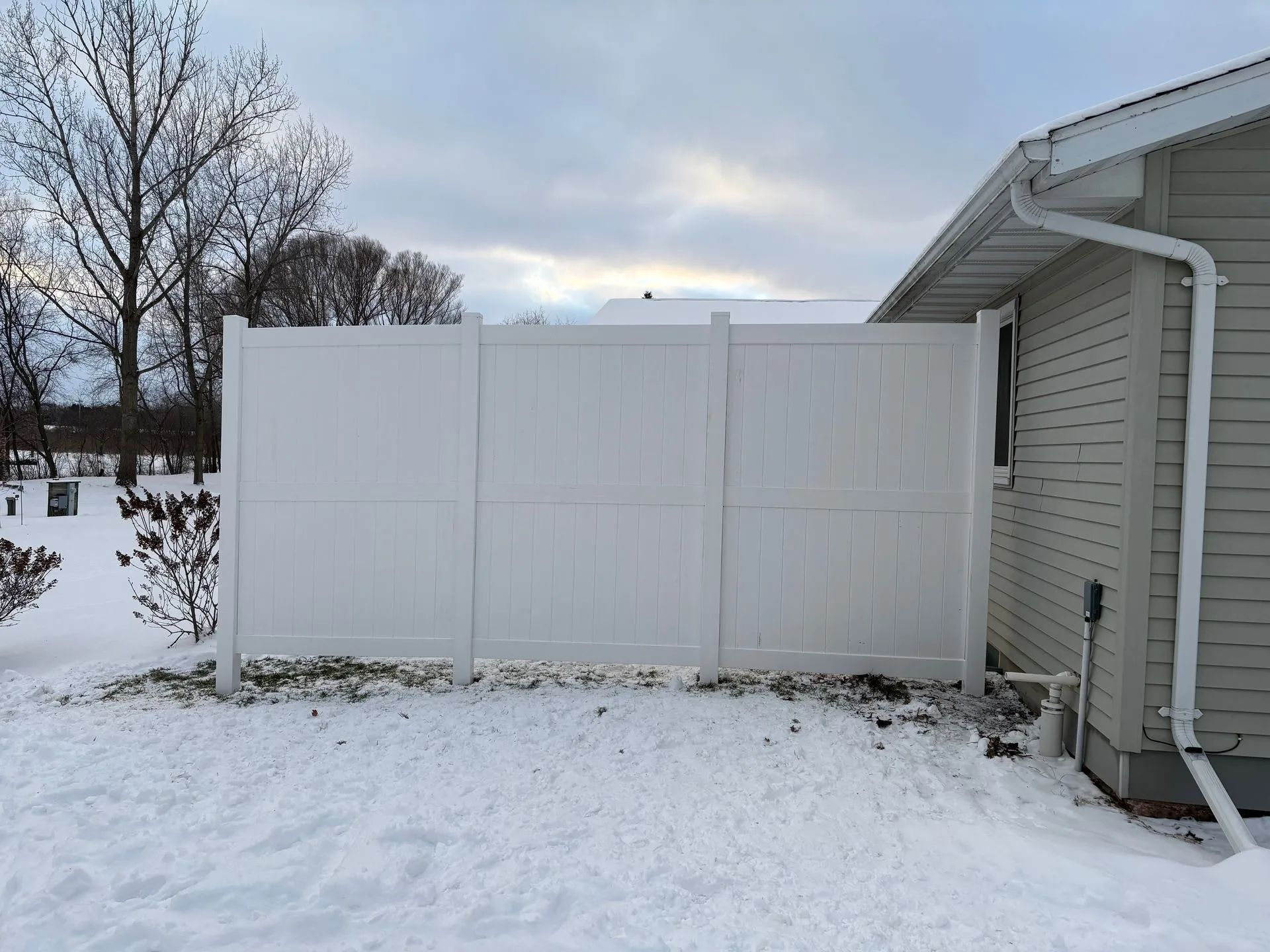 White vinyl fence next to a house with snow on the ground; cloudy sky.